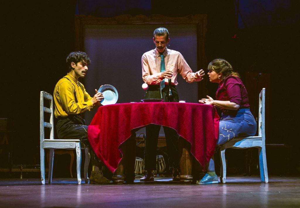 Tres personas sentadas alrededor de mesa roja en escenario de teatro durante conversación o debate sobre la humanidad