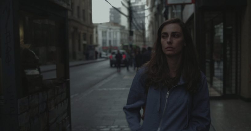 Mujer joven en calle urbana al atardecer, viste chaqueta azul oscuro y mira hacia la cámara con expresión serena