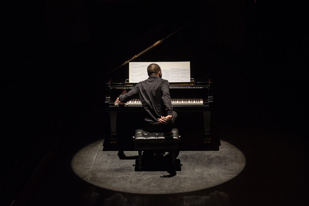 Wilhem Latchoumia tocando piano de cola en escenario iluminado con fondo oscuro y proyección de video