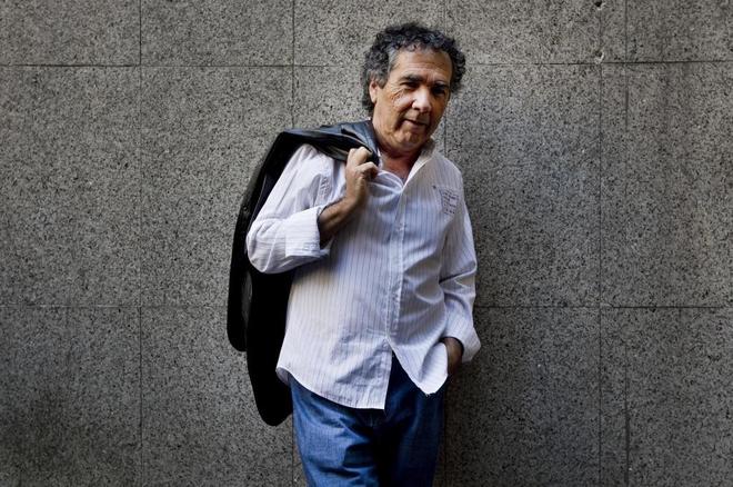 Hernán Rivera Letelier, escritor chileno, sostiene su chaqueta negra sobre el hombro frente a pared de hormigón gris