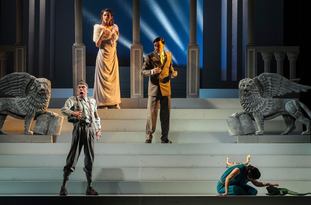 Escena teatral con actores en escenario azul: figura en primer plano, personajes en túnicas doradas al fondo y actor
