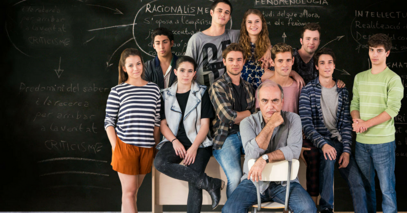 Grupo de estudiantes y profesor frente a pizarra con escritura, escena de la serie que muestra ambiente educativo
