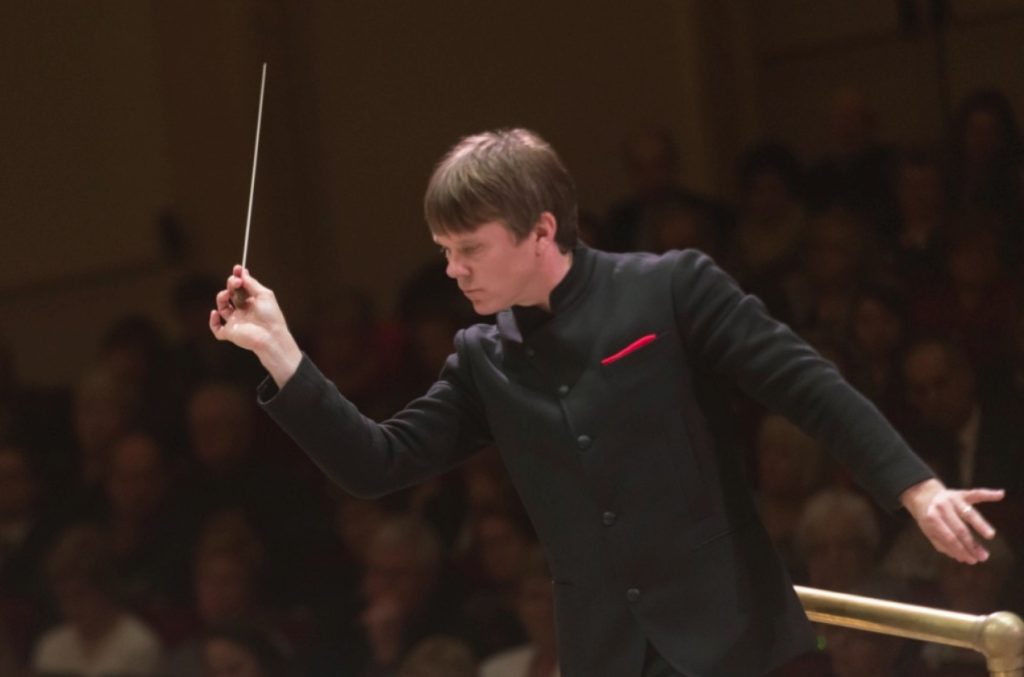 Director alemán con batuta dirigiendo orquesta sinfónica, vistiendo traje negro formal con corbata roja