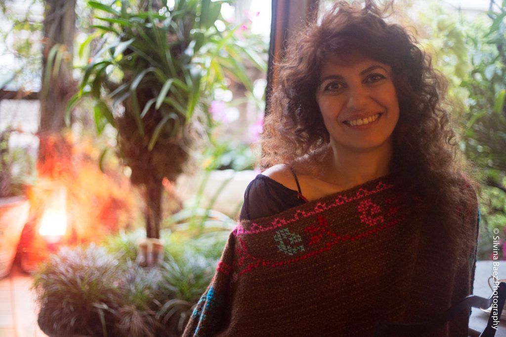 La entrevista patafísica: Ana Arzoumanian, entre el sigilo y la corrida voraz Mujer sonriente con cabello rizado oscuro, vistiendo suéter granate, rodeada de plantas verdes en interior iluminado