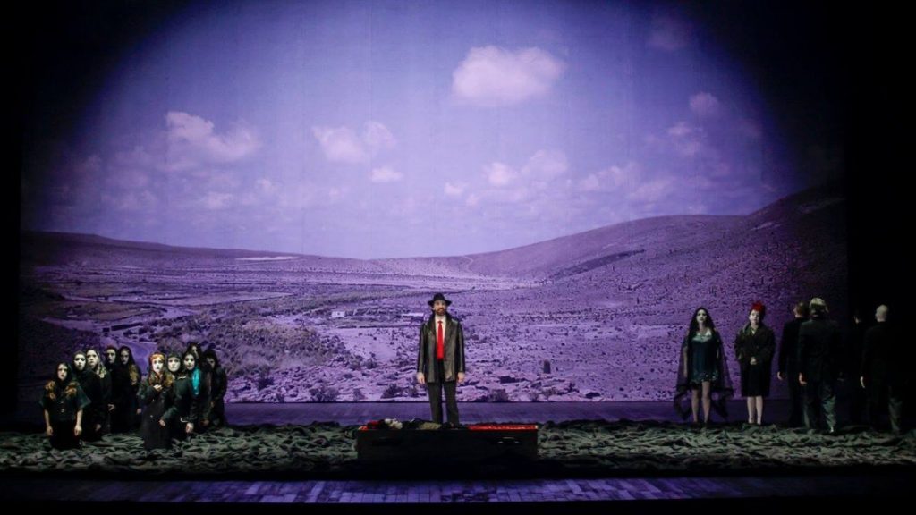 Escena teatral con figura central bajo cielo púrpura, personajes en silhueta a ambos lados en montaña desértica