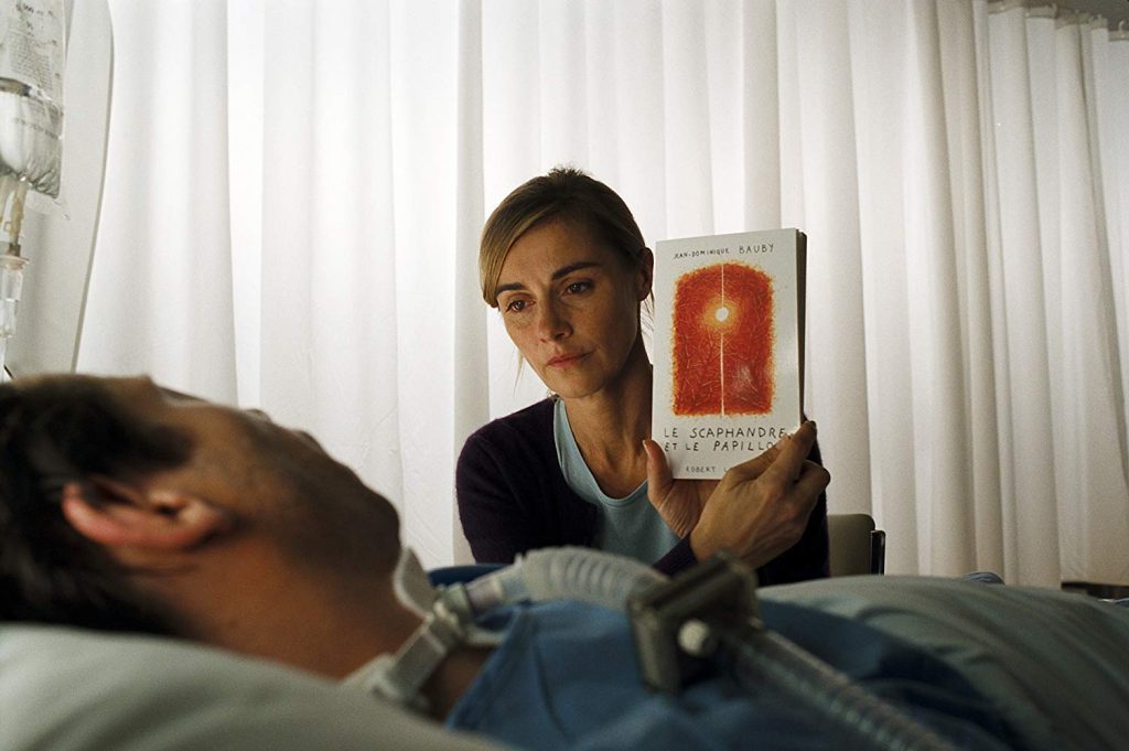 Mujer sostiene un libro de portada roja mientras lee acostada en una cama, luz natural de fondo