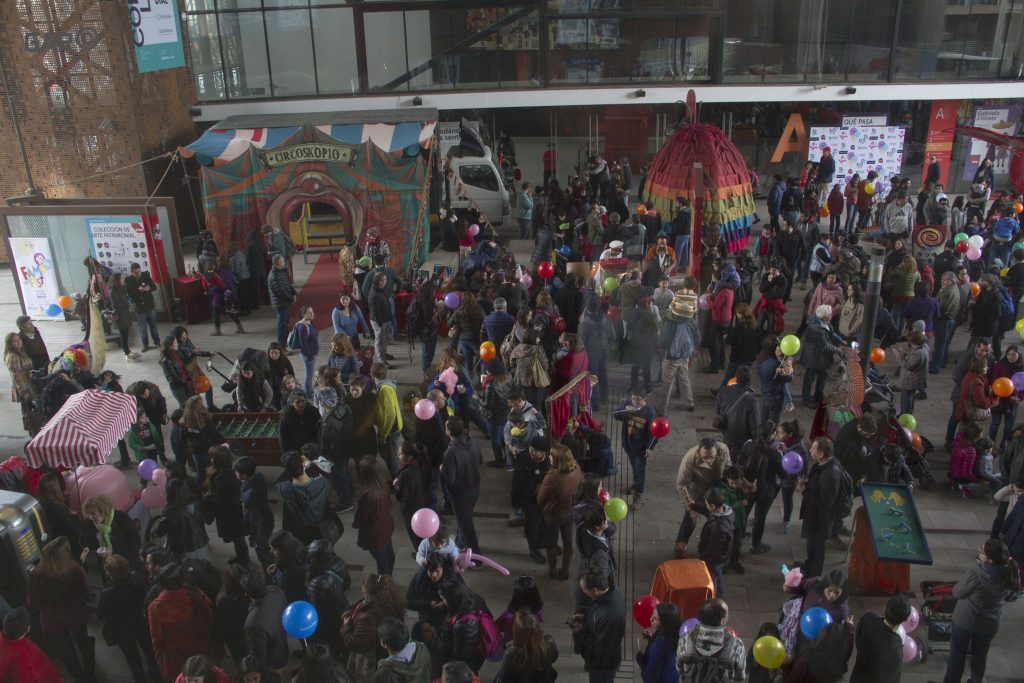 Multitud de personas en una plaza urbana durante festival de teatro familiar, con tiendas y estructuras de eventos