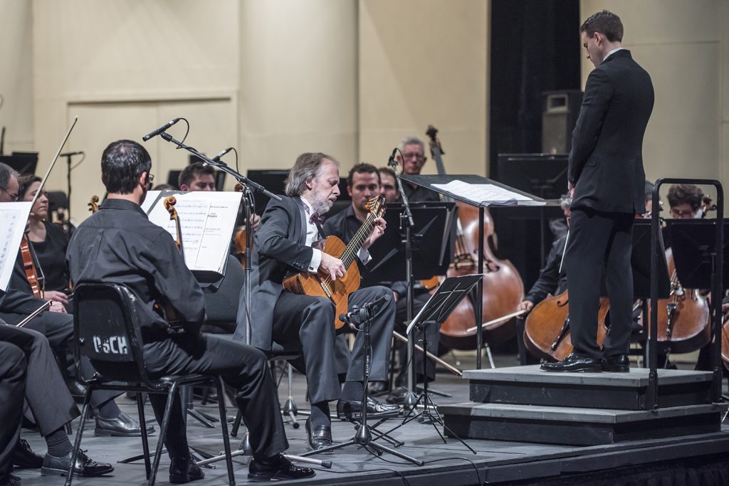 Orquesta de cámara en concierto, director en podio, músicos con instrumentos de cuerda y vientos en escenario