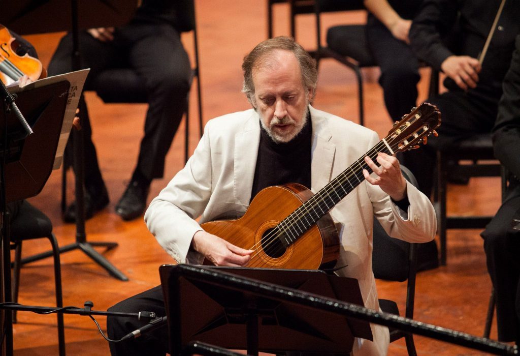 El destacado guitarrista Luis Orlandini reestrenará una importante obra chilena para cuerdas junto a la Orquesta Sinfónica Nacional Guitarrista Luis Orlandini toca guitarra acústica en concierto con orquesta sinfónica al fondo