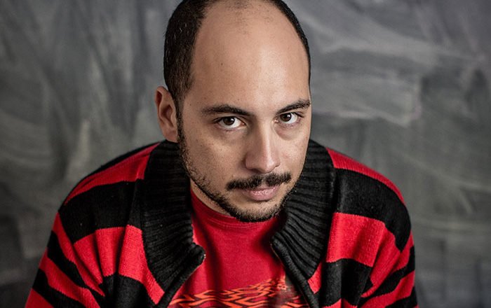 Nicolás López, cineasta chileno, retratado con camisa a cuadros roja y negra frente a fondo gris