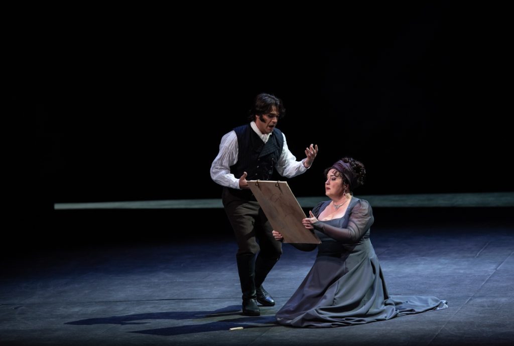 Escena de la ópera Tosca: hombre en traje y chaleco sostiene a mujer arrodillada en vestido gris sobre escenario oscuro