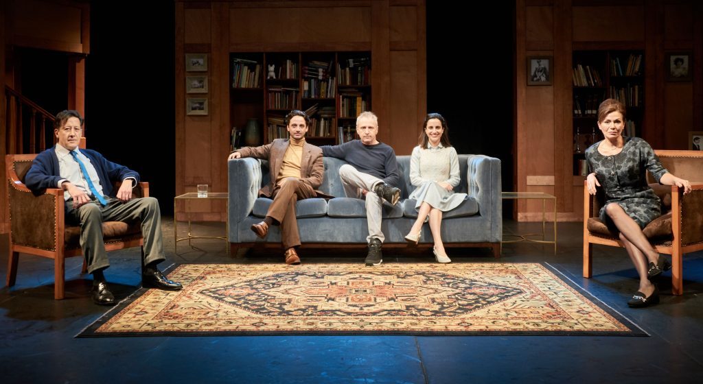 Cinco actores en sala de estar teatral con sofá central, alfombra y libreros de fondo, escena de obra dramática