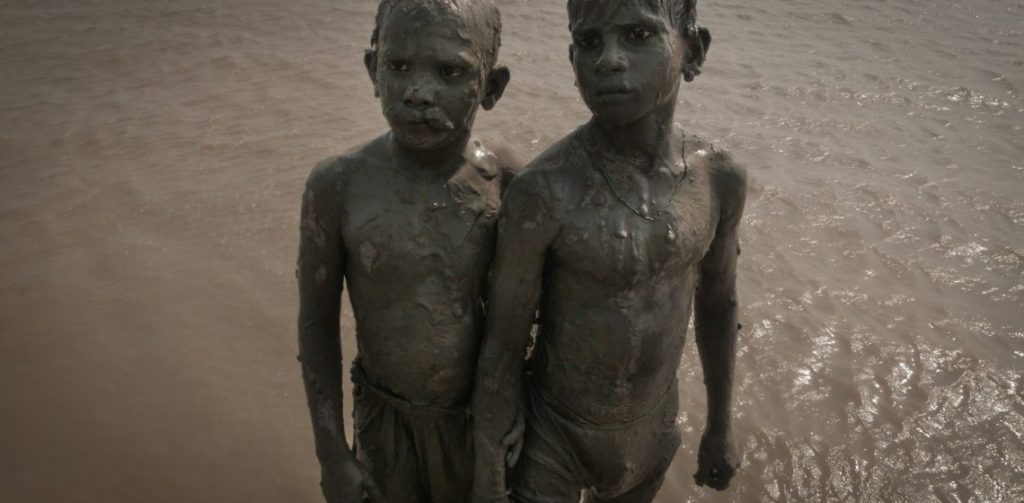 Dos niños cubiertos de lodo en agua turbia, mirando a cámara con expresión seria en ambiente natural