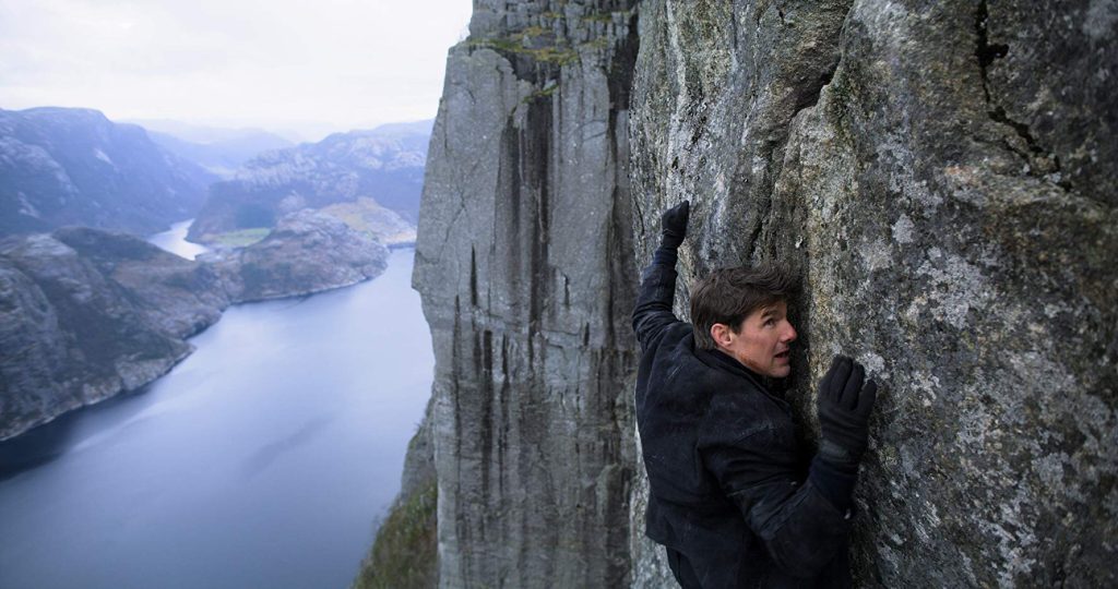 "Misión imposible: Repercusión": Las tensiones increíbles de la existencia Hombre escalando acantilado vertical de roca gris sobre lago alpino con montañas nevadas al fondo