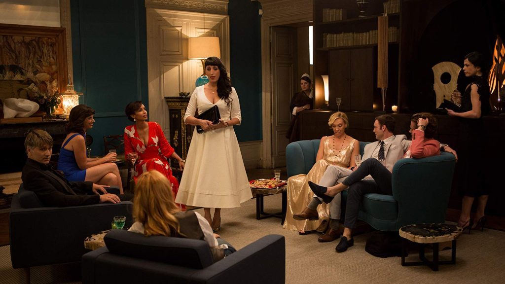 Escena de película con mujer en vestido blanco de pie en centro de sala mientras grupo de personas la observa sentadas