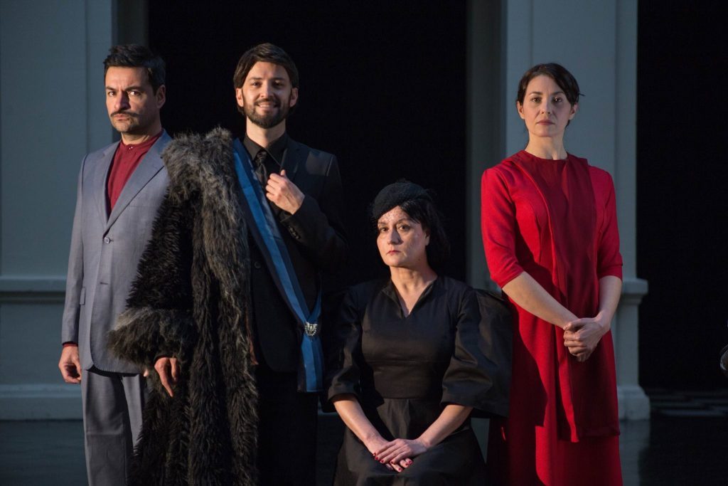 Cuatro actores en escena teatral: hombre con abrigo gris a la izquierda, otro con capa azul, mujer de cabello oscuro