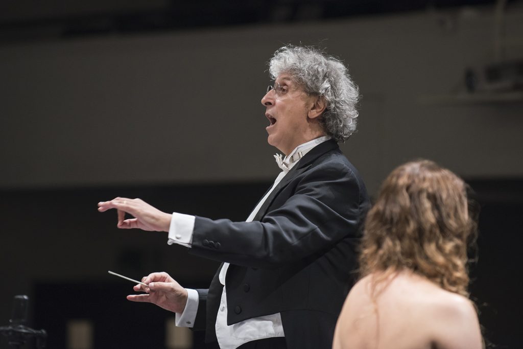 Director de orquesta con cabello blanco gesticulando frente a músicos durante ensayo o concierto de la Orquesta