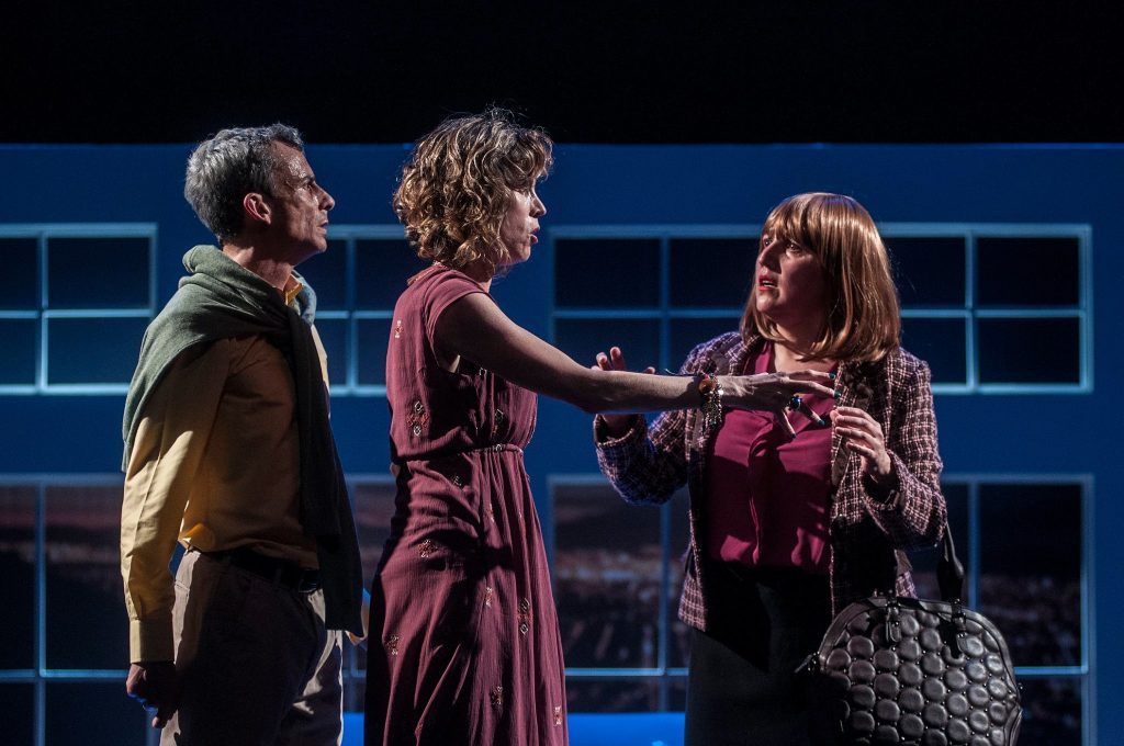 "Noche mapuche", de Marcelo Leonart: El infortunio histórico del placer carnal Tres actores en escena teatral: hombre con chaqueta amarilla, mujer en vestido burdeos y otra mujer con bolso