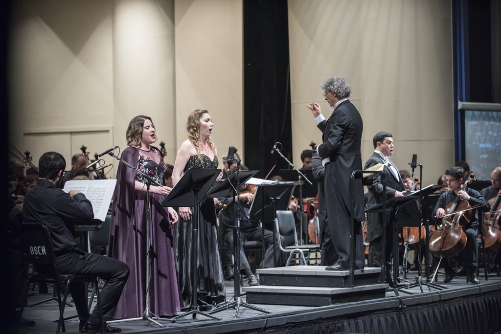 Orquesta y cantantes líricos en escena durante concierto clásico, con director frente a músicos en auditorio