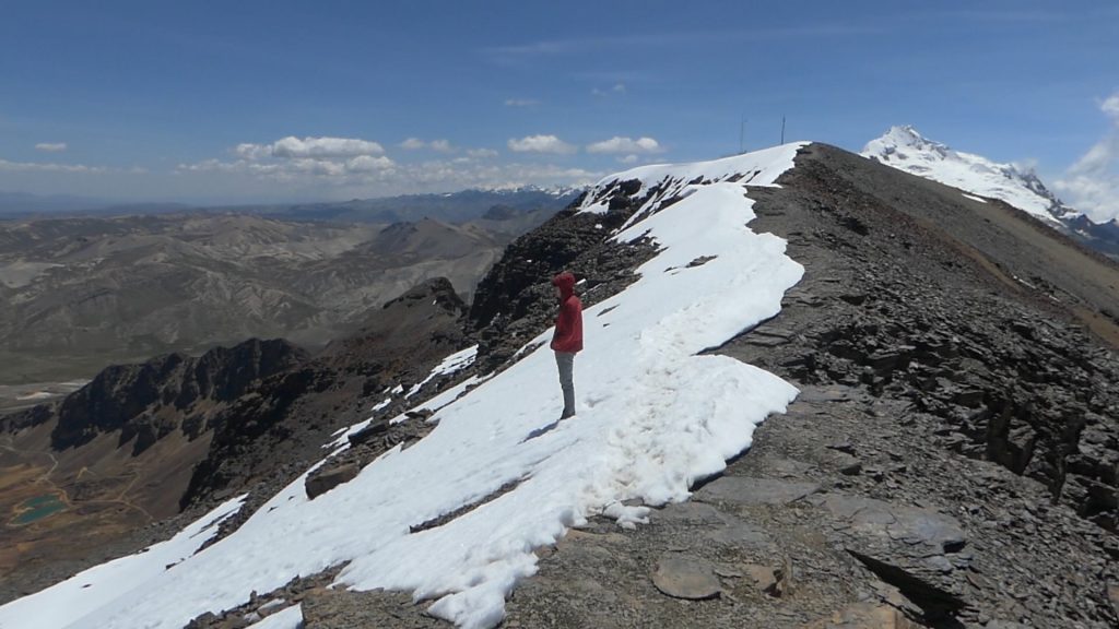 Montañista en pendiente nevada de alta montaña, caminando hacia cumbre nevada bajo cielo despejado con valles al fondo