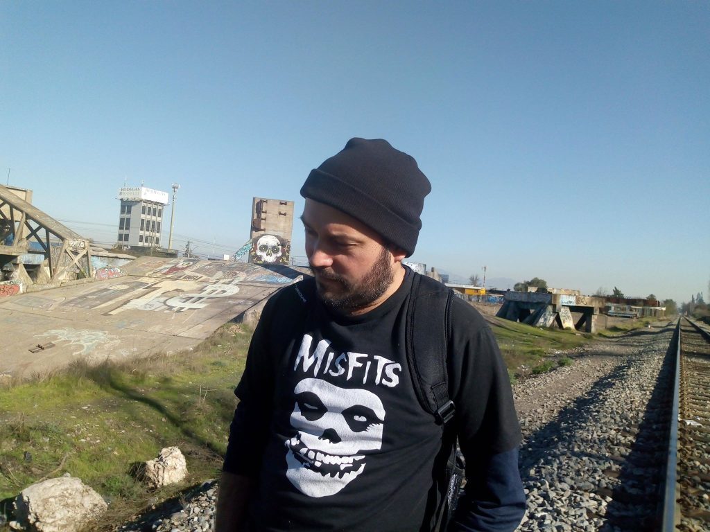 Andrés Torres Meza vistiendo remera con logo Misfits, gorro negro, junto a vías de tren y construcción industrial