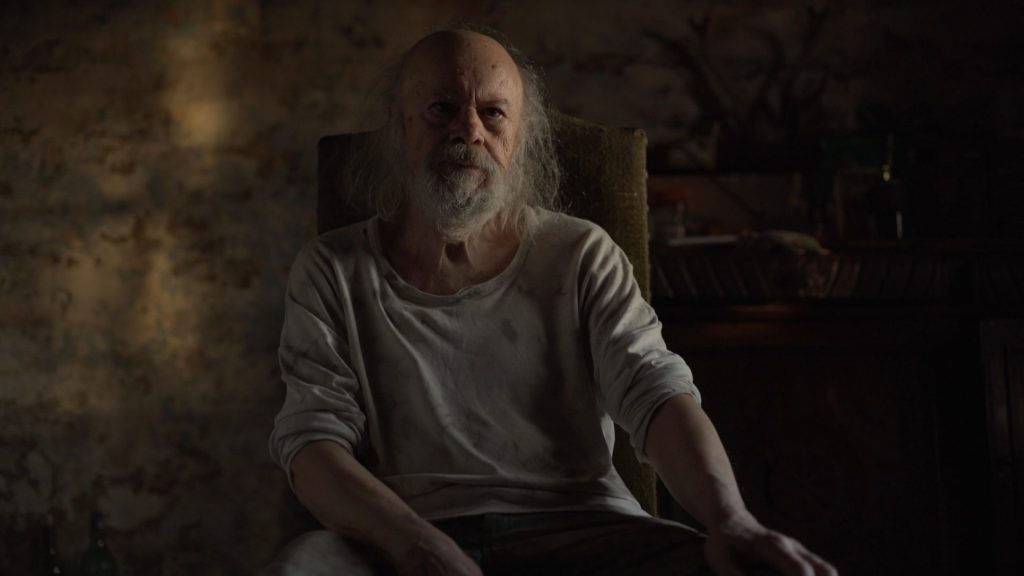 Hombre mayor con barba blanca en habitación oscura, escena del documental El espanto sobre sistemas de salud públicos