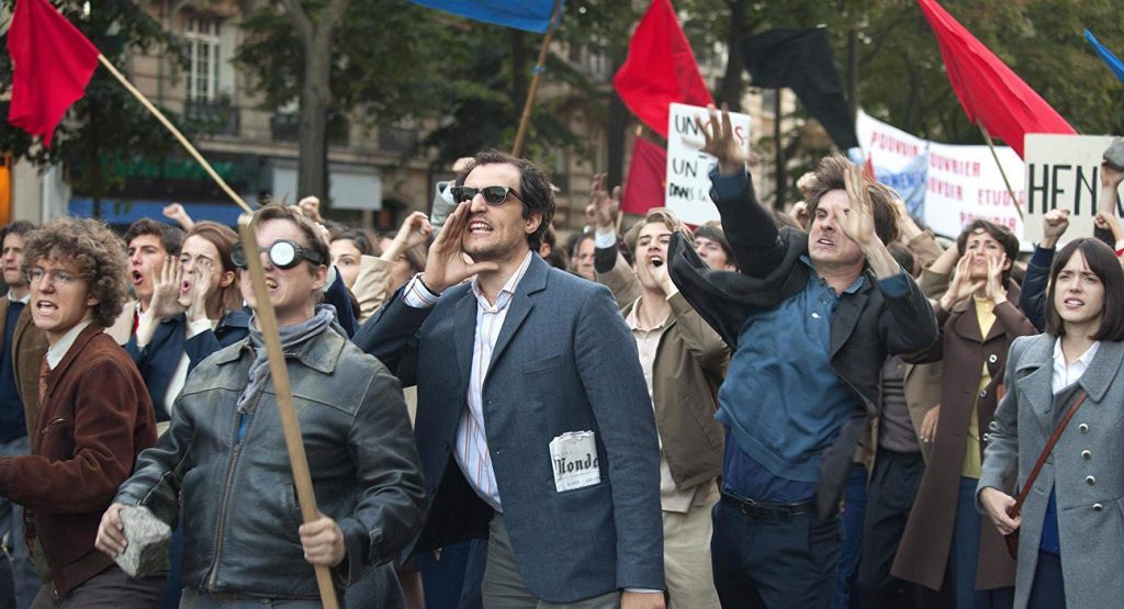Manifestación callejera con personas portando banderas rojas, hombres en traje de época y multitud de asistentes