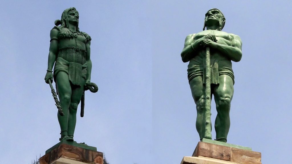 Dos estatuas de bronce de guerreros indígenas en posición de pie sobre pedestales de piedra, vistas de frente