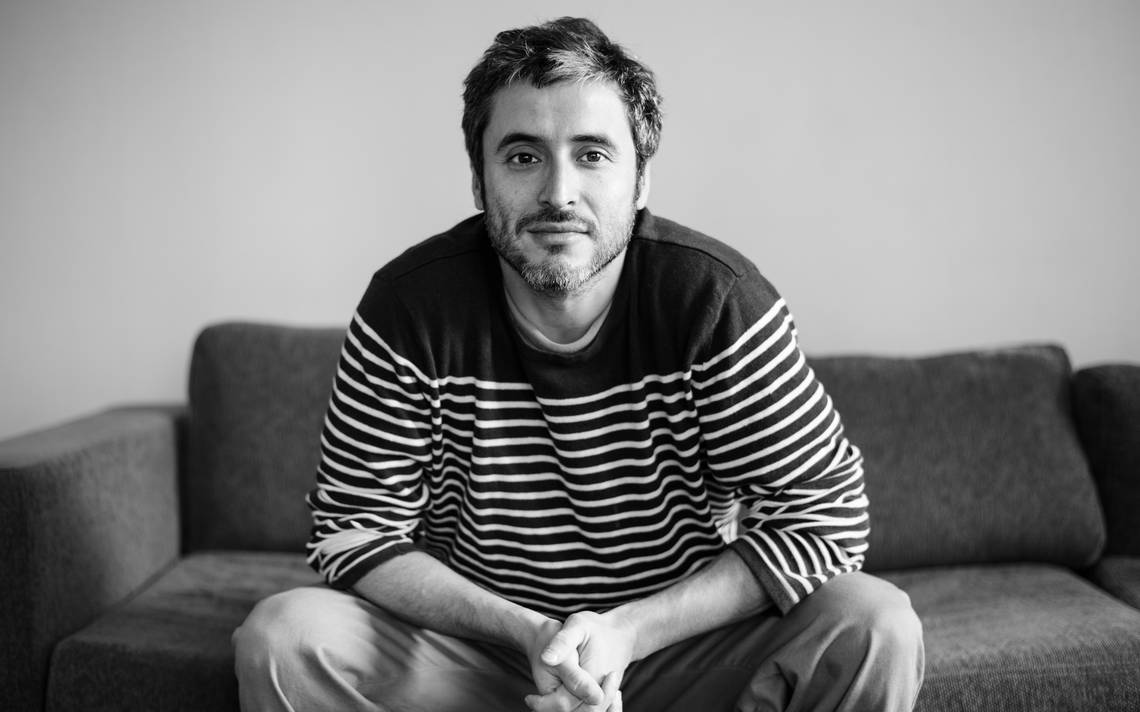 Javier Rebolledo sentado en sofá gris, vistiendo suéter a rayas, mirada directa a cámara en fotografía blanco y negro