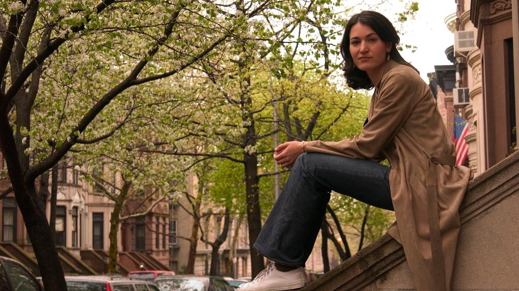 Llega una mujer y dice: Nicole Krauss (una crónica de Andrea Jeftanovic) Nicole Krauss sentada en una barandilla de piedra en una calle arbolada de edificios históricos
