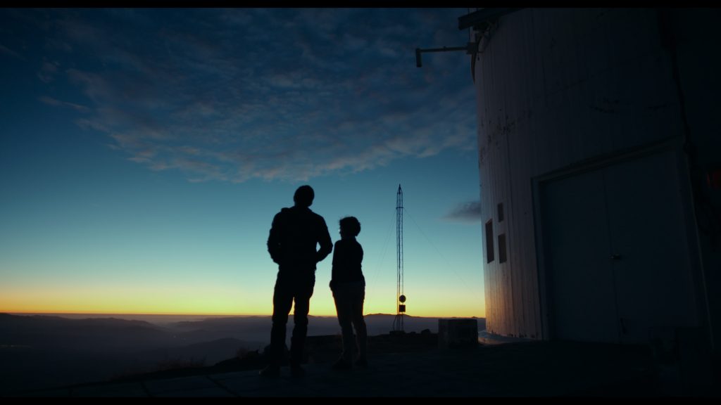 Dos siluetas observan el cielo nocturno junto a un observatorio astronómico al atardecer en el desierto de Atacama