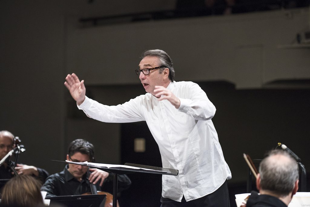 Director de orquesta Leonid Grin gesticula con ambas manos durante ensayo o concierto, músicos al fondo