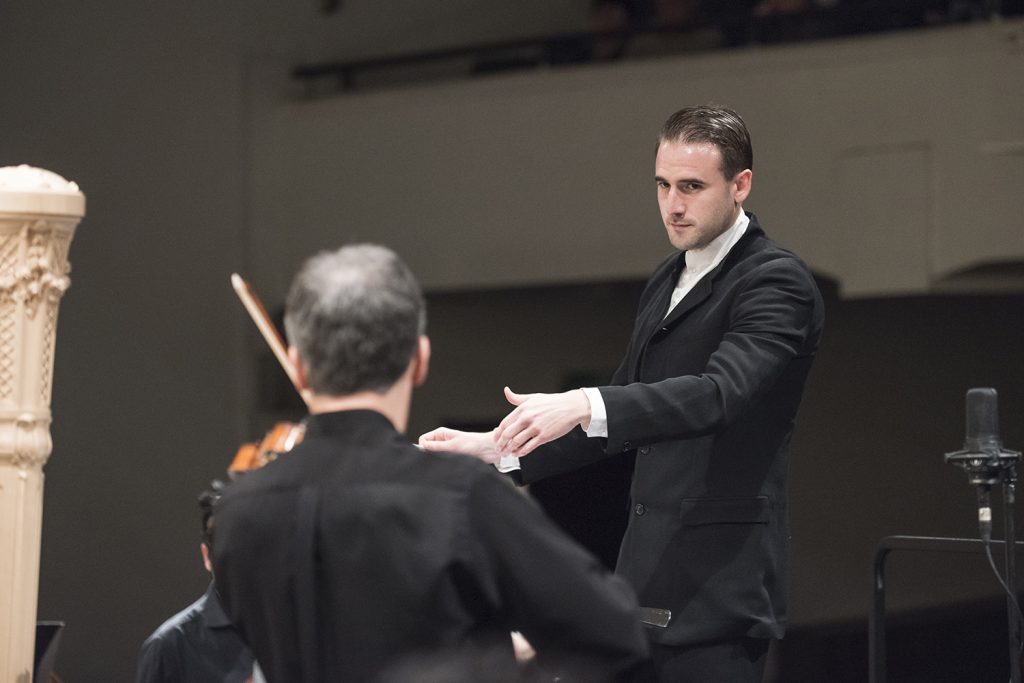 Concierto 14 del Ceac de la Universidad de Chile: Una versión dubitativa de la "Nuevo mundo" de Dvorak Director de orquesta gesticulando mientras dirige a músicos durante concierto en sala de conciertos
