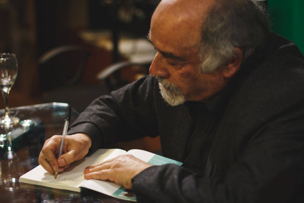 Cuentos de "Bucear en su alma", de Juan Mihovilovich: Un tributo a la literatura fantástica de Chile Hombre mayor escribiendo en un cuaderno durante un acto literario, pluma en mano y vaso de agua al fondo