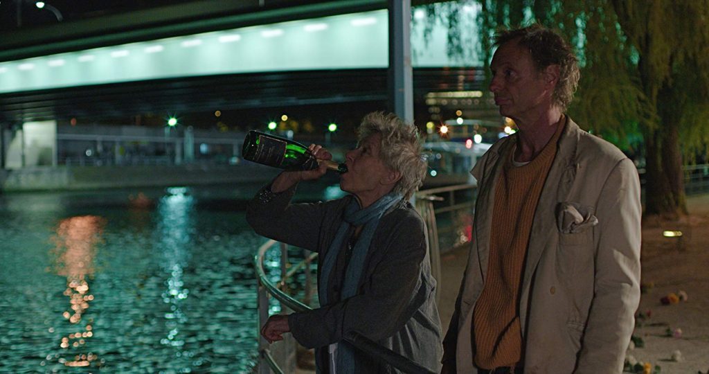 Dos personas de pie junto a un río nocturno iluminado, con arquitectura urbana de fondo