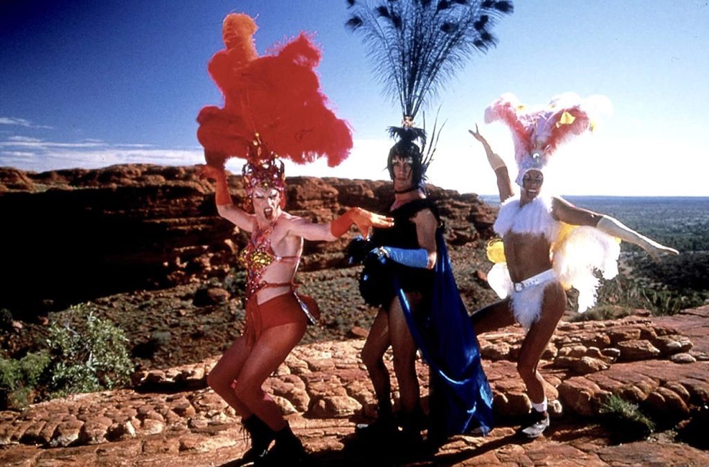 Tres performers en trajes coloridos bailan en un paisaje desértico con montañas al fondo bajo el cielo azul