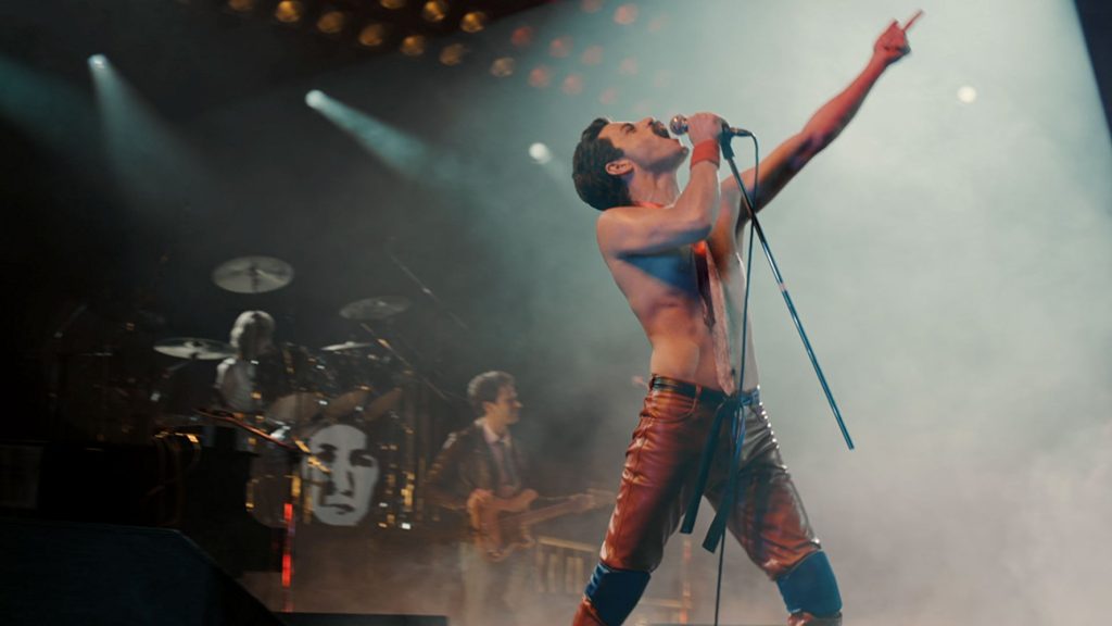Freddie Mercury cantando en el escenario con el brazo levantado, micrófono en mano, rodeado de luces y la banda en vivo