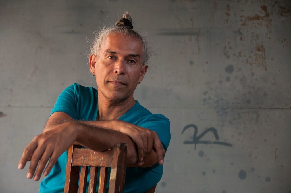 Ciro Beltrán, hombre con cabello gris recogido, viste camisa azul y apoya los brazos sobre una silla de madera