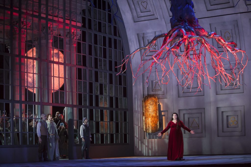 Escena de la ópera Norma con actriz en vestido rojo ante arquitectura neoclásica e iluminación teatral roja