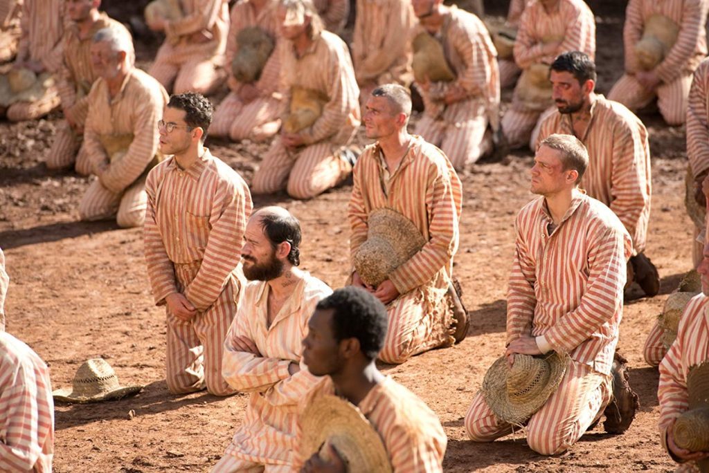 "Papillon, la gran fuga", de Michael Noer: Una nueva versión de un clásico sobre la cárcel Prisioneros en uniforme rayado arrodillados en patio de tierra de prisión tropical con guardias vigilando