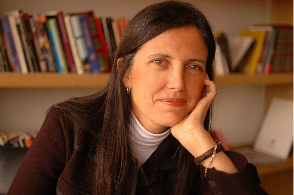 Claudia Piñeiro, escritora argentina, apoyada en su mano con expresión reflexiva frente a estantería con libros