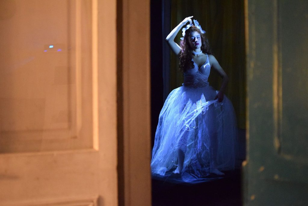 Mujer en vestido azul translúcido de noche, con mano en cabeza, en escena teatral iluminada con tonos azules