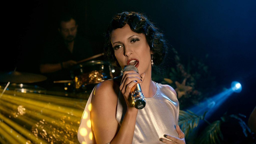 Mujer cantante con micrófono en escena, iluminación azul y amarilla, músico de fondo en penumbra