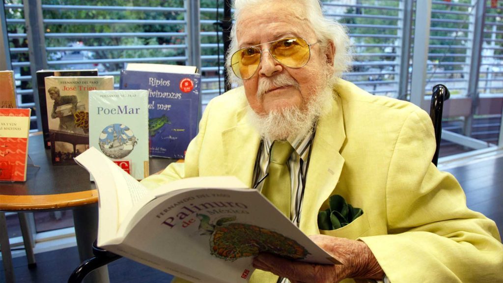 Fernando del Paso sostiene una revista en su oficina con vista a la ciudad, vistiendo blazer amarillo