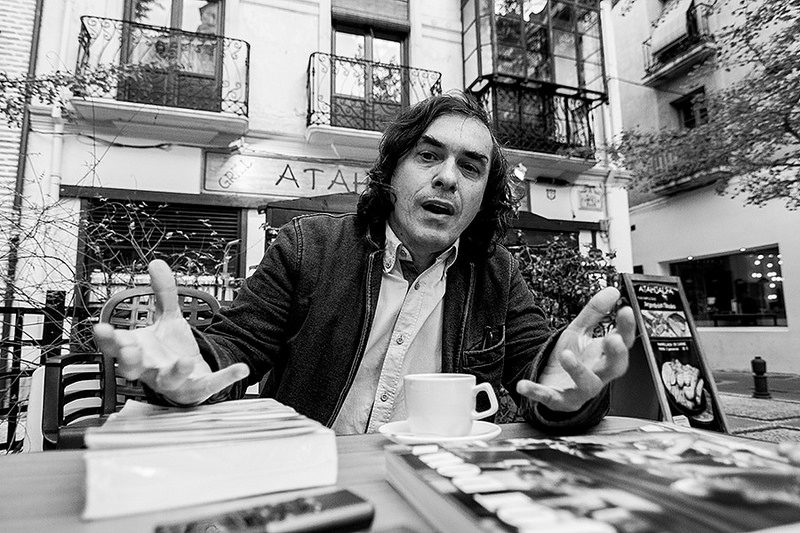Mircea Cărtărescu gesticula mientras habla en una mesa, rodeado de libros y materiales de lectura en un espacio urbano