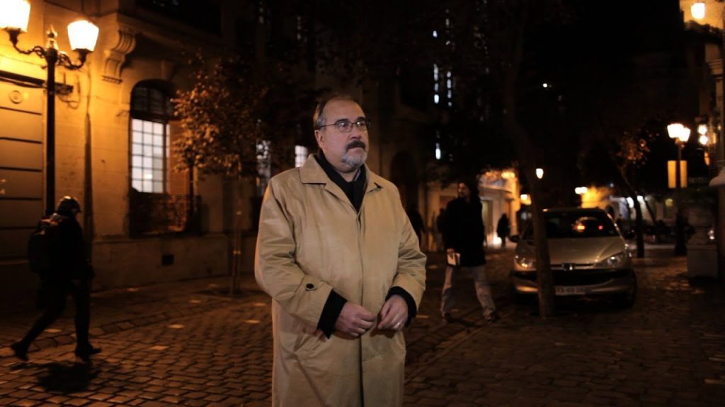Ramón Díaz Eterovic en la calle de noche, con abrigo beige y gafas, en un entorno urbano iluminado