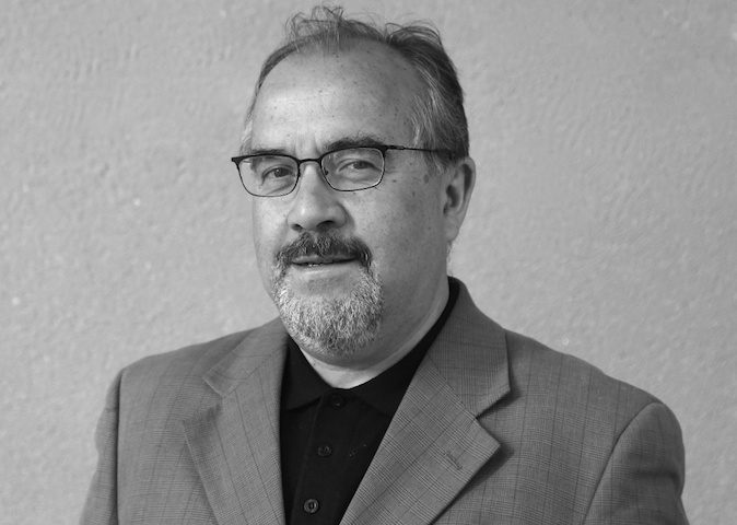 Ramón Díaz Eterovic, escritor chileno, en fotografía de perfil con gafas y traje gris sobre fondo neutro