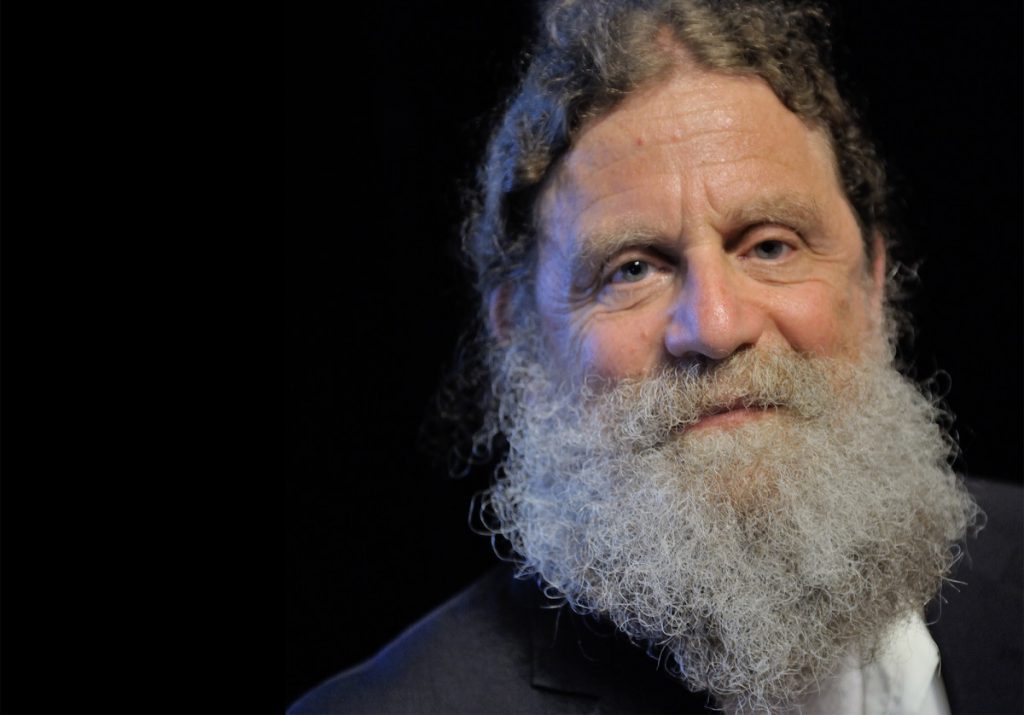 Hombre mayor con barba gris abundante, cabello gris oscuro, fondo negro. Retrato de Robert Sapolsky