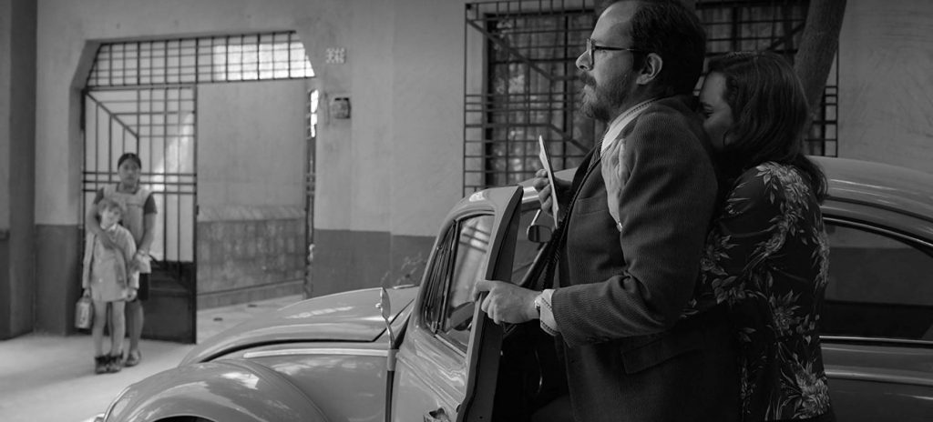 Escena en blanco y negro de interiores de una casa, mujer indígena en primer plano observa a dos personas conversando