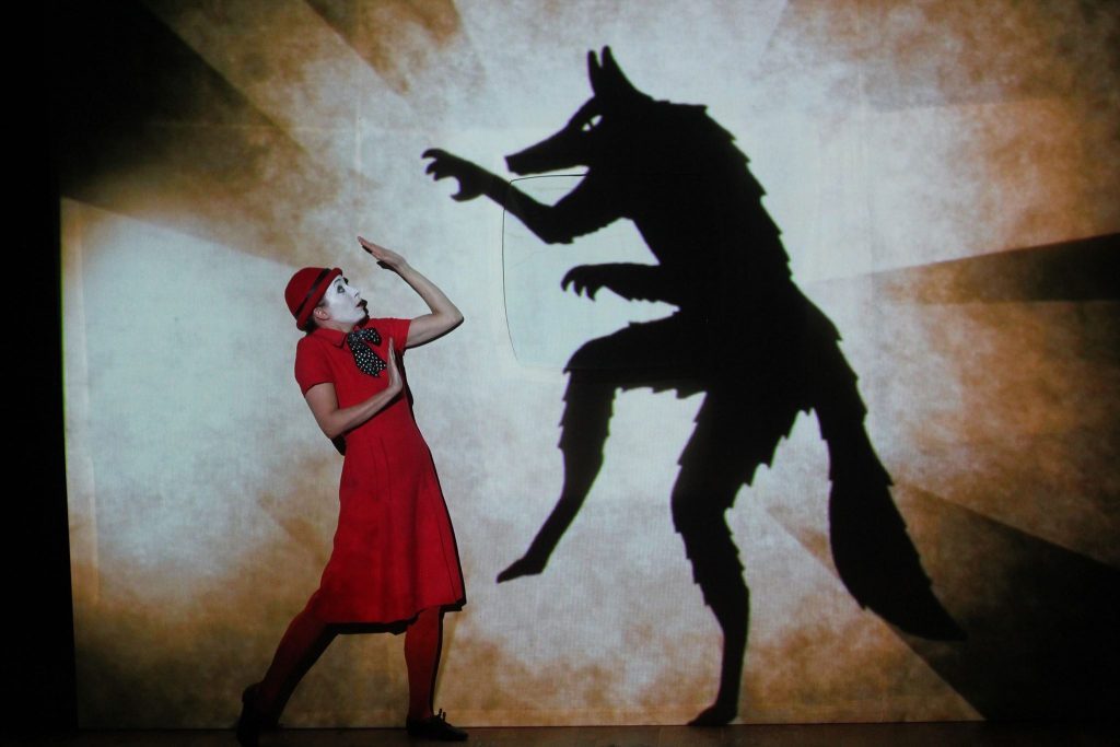 "The Animals And Children Took To The Streets" de la Compañía 1927, en Chile: Un espectáculo fenomenal Mujer en vestido rojo se defiende de silueta de criatura oscura en teatro de sombras, escena dramática de contraste luz